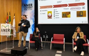 Paulo Henrique Ferreira, da Barões, fala em um microfone no palco ao lado de Sabrina Passos e Ruth Reis. Atrás deles, há uma tela grande que mostra uma linha do tempo de transição da mídia e um banner que diz ANER Summit 2026. Três cadeiras vazias também são visíveis.