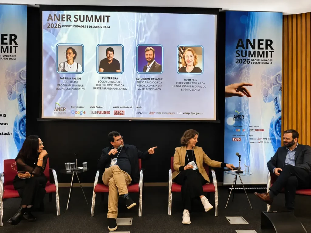 Cinco palestrantes sentados em um palco durante o ANER Summit 2026. Atrás deles, há uma tela exibindo seus nomes, fotos e títulos, juntamente com a marca do evento e os logotipos dos patrocinadores. Quatro painelistas estão conversando.