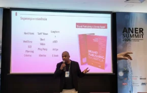 Um homem fala em um microfone em frente a uma tela de apresentação que exibe um livro intitulado Brand Publishing na Prática e slides sobre tipos de conteúdo. O banner do evento diz ANER Summit.