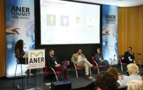 Cinco pessoas participam de um painel de discussão no ANER Summit 2026. Quatro estão sentadas no palco e uma está em um púlpito. Uma tela grande atrás deles exibe informações sobre o palestrante. Os membros da plateia observam atentamente.