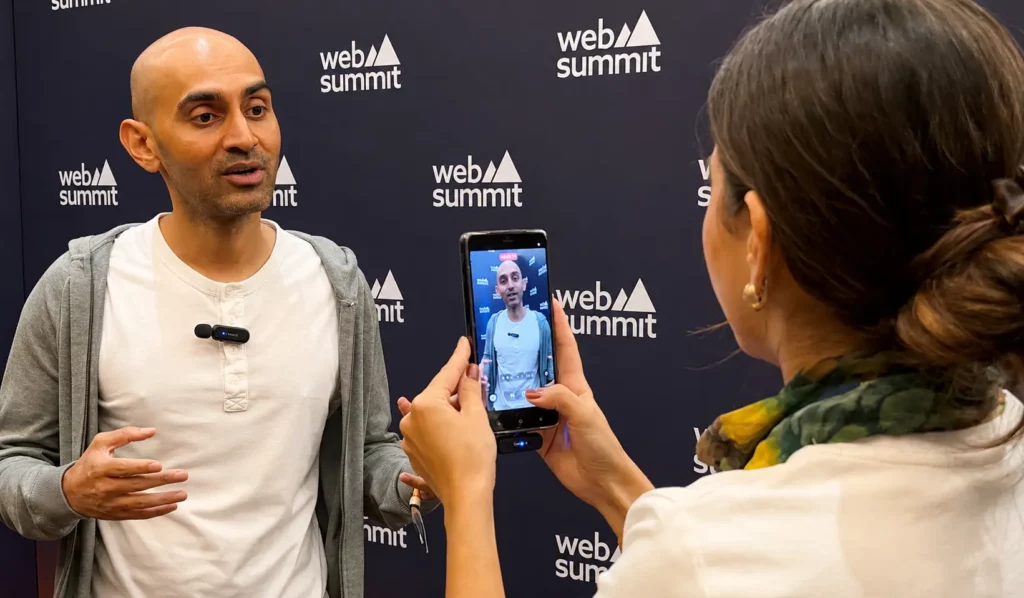 Um homem fala em frente a um cenário do Web Summit enquanto uma mulher o filma com seu smartphone. A tela do telefone mostra o homem sendo gravado enquanto ele faz gestos com as mãos.