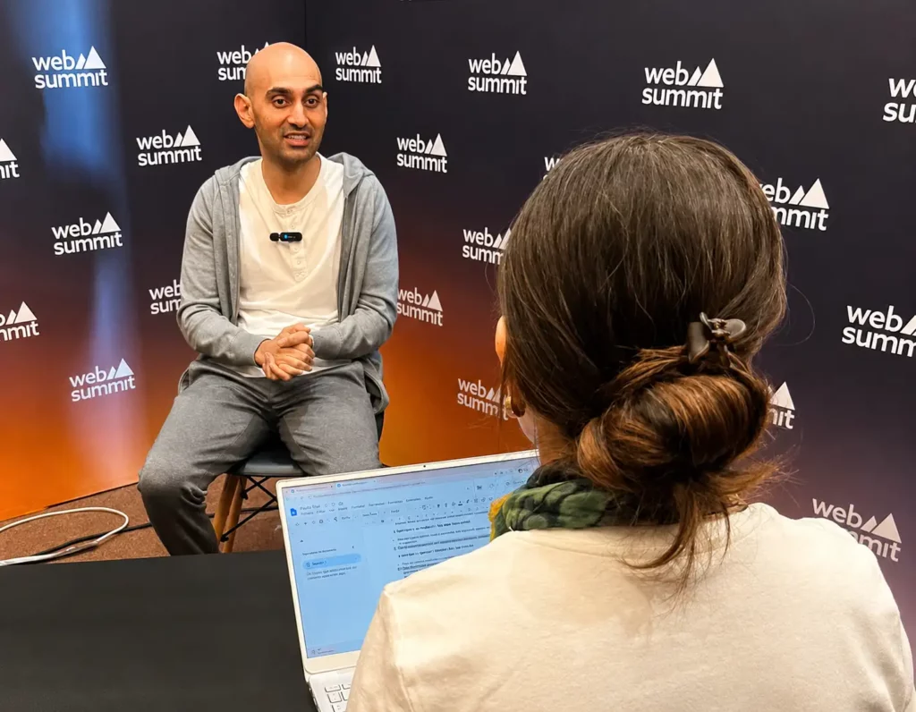 Um homem está sentado em um banco, falando com uma mulher que está digitando em um laptop durante uma entrevista. Ambos estão em frente a um pano de fundo preto com logotipos do Web Summit.