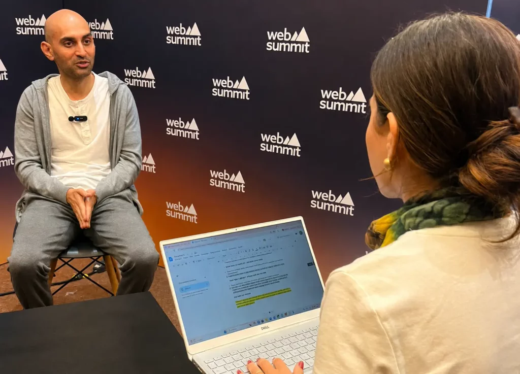 Um homem em roupas casuais senta-se para uma entrevista no Web Summit enquanto uma mulher com um laptop faz anotações, de frente para ele. O logotipo do evento está visível no pano de fundo atrás deles.