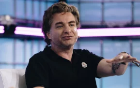 Uma pessoa de cabelo castanho, vestindo uma camisa polo preta, está sentada e falando com uma mão estendida. É possível ver uma tatuagem no antebraço dela, e o fundo apresenta luzes roxas e painéis de vidro.
