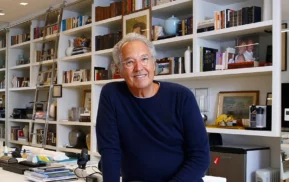 Um senhor de cabelos grisalhos e óculos, vestindo um suéter azul-marinho, sorri enquanto está sentado em um escritório moderno, tendo ao fundo prateleiras brancas cheias de livros, fotos emolduradas e objetos decorativos.