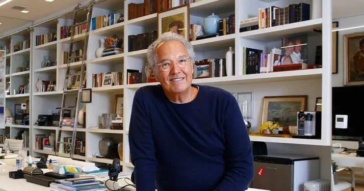 Um senhor de cabelos grisalhos e óculos, vestindo um suéter azul-marinho, sorri enquanto está sentado em um escritório moderno, tendo ao fundo prateleiras brancas cheias de livros, fotos emolduradas e objetos decorativos.