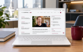 Um laptop sobre uma mesa exibe um site com um artigo em que aparece um homem sorrindo. Perto dele, há uma caneca vermelha, uma caneta e cadernos fechados. Ao fundo, há estantes de livros e uma planta.