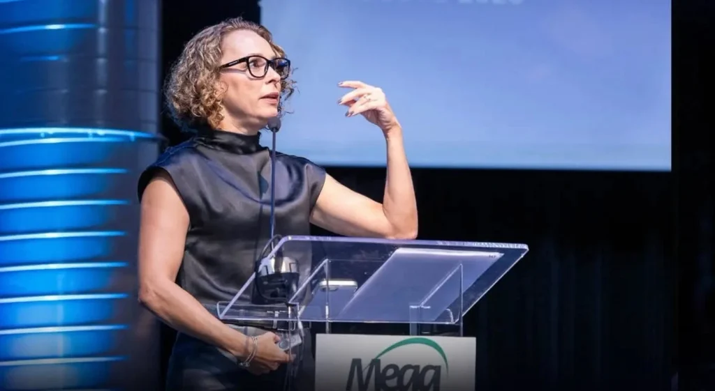 Uma pessoa com cabelos cacheados e óculos fala em um pódio transparente com o logotipo da Mega, gesticulando com uma mão, tendo como pano de fundo uma luz azul.
