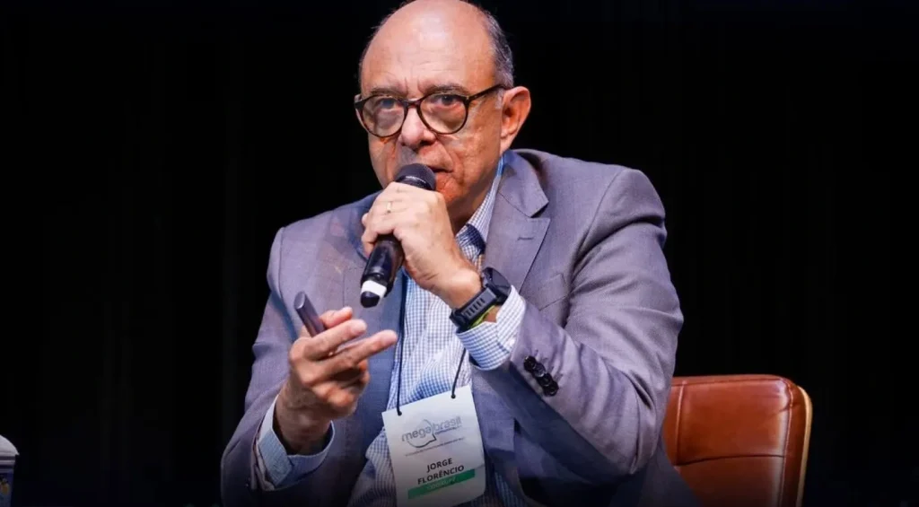 Um homem de terno cinza e óculos fala ao microfone enquanto está sentado, segurando um aparelho na outra mão. Ele usa um crachá e tem uma expressão séria, com um fundo escuro atrás dele.