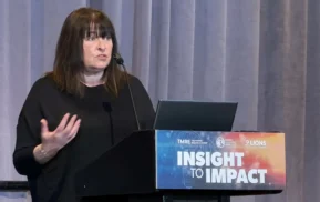 Uma mulher vestindo uma camisa preta discursa em um pódio com a inscrição Insight to Impact em uma conferência da TMRE.