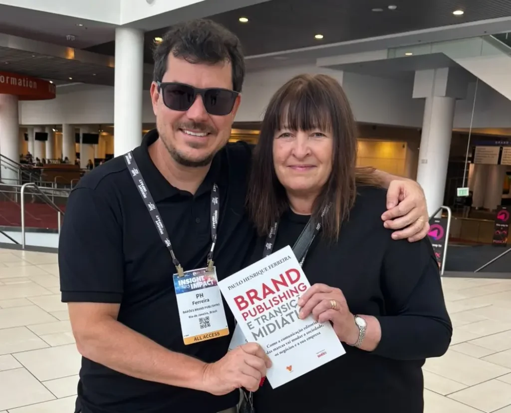 Um homem usando óculos escuros e uma mulher segurando um exemplar do livro *Brand Publishing e Transição Midiática* posam juntos em um amplo saguão.